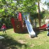 Bambini giocano in un parco con scivolo a forma di nave e altalene.