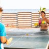 Due persone giocano a ping-pong su una spiaggia, con il mare sullo sfondo.