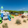 Parco giochi sabbioso con scivoli verdi e blu, torri e tema di castello sotto un cielo azzurro.