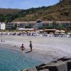 Persone giocano in acqua su una spiaggia di ciottoli, case e colline sullo sfondo.